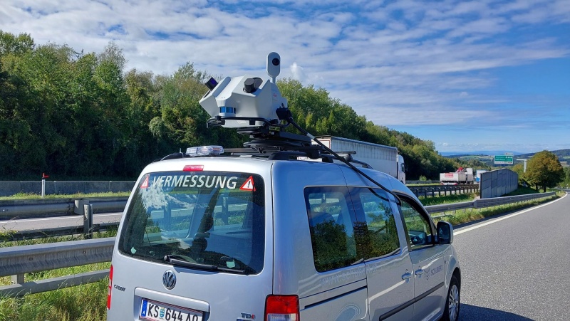 Ein Auto fährt eine Straße entlang, links ist Wald, am Autodach ist ein Laserscanner installiert