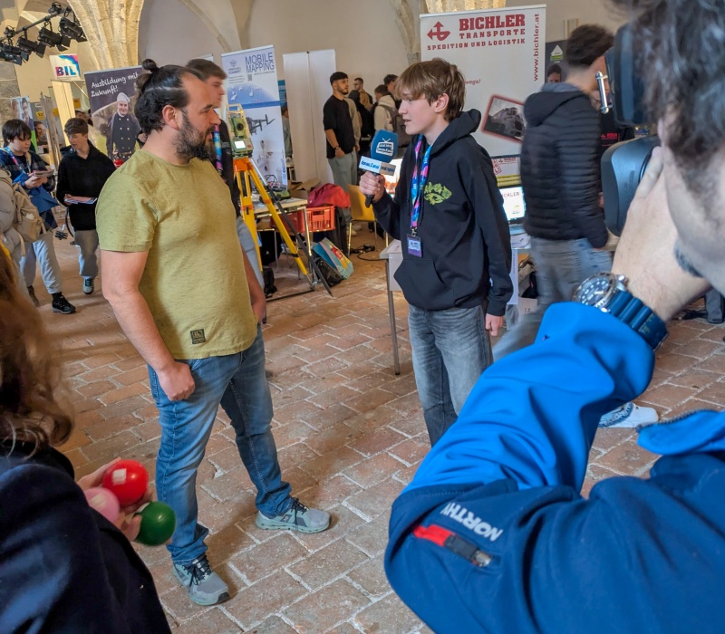 Ein Jugendlicher im Lehrlingsalter führt ein Interview mit einem Mann vor einem Messestand