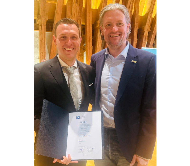 2 Herren in Anzug stehen nebeneinander, der j&uuml;ngere der beiden h&auml;lt eine Urkunde