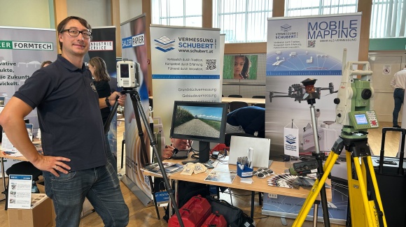 1 Mann steht neben einem Messestand mit technischen Geräten und Werbefolder, dahinter stehen Roll-Ups,